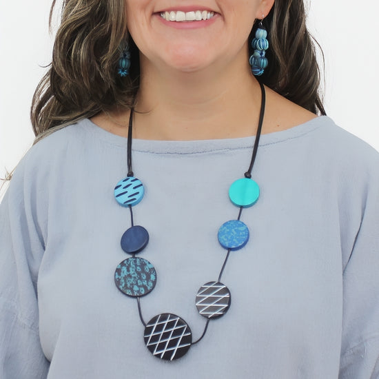 A woman wearing a multi-piece necklace with circular beads in various patterns and colors, including blue, teal, and black-and-white geometric designs. She is also wearing matching blue patterned drop earrings and a light blue-gray top against a plain white background.