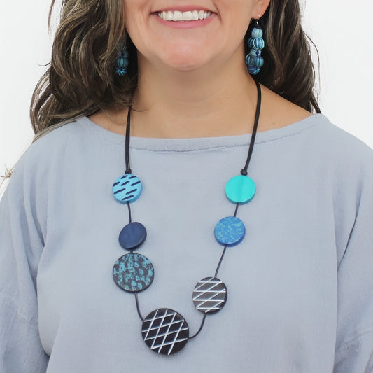 A woman wearing a multi-piece necklace with circular beads in various patterns and colors, including blue, teal, and black-and-white geometric designs. She is also wearing matching blue patterned drop earrings and a light blue-gray top against a plain white background.