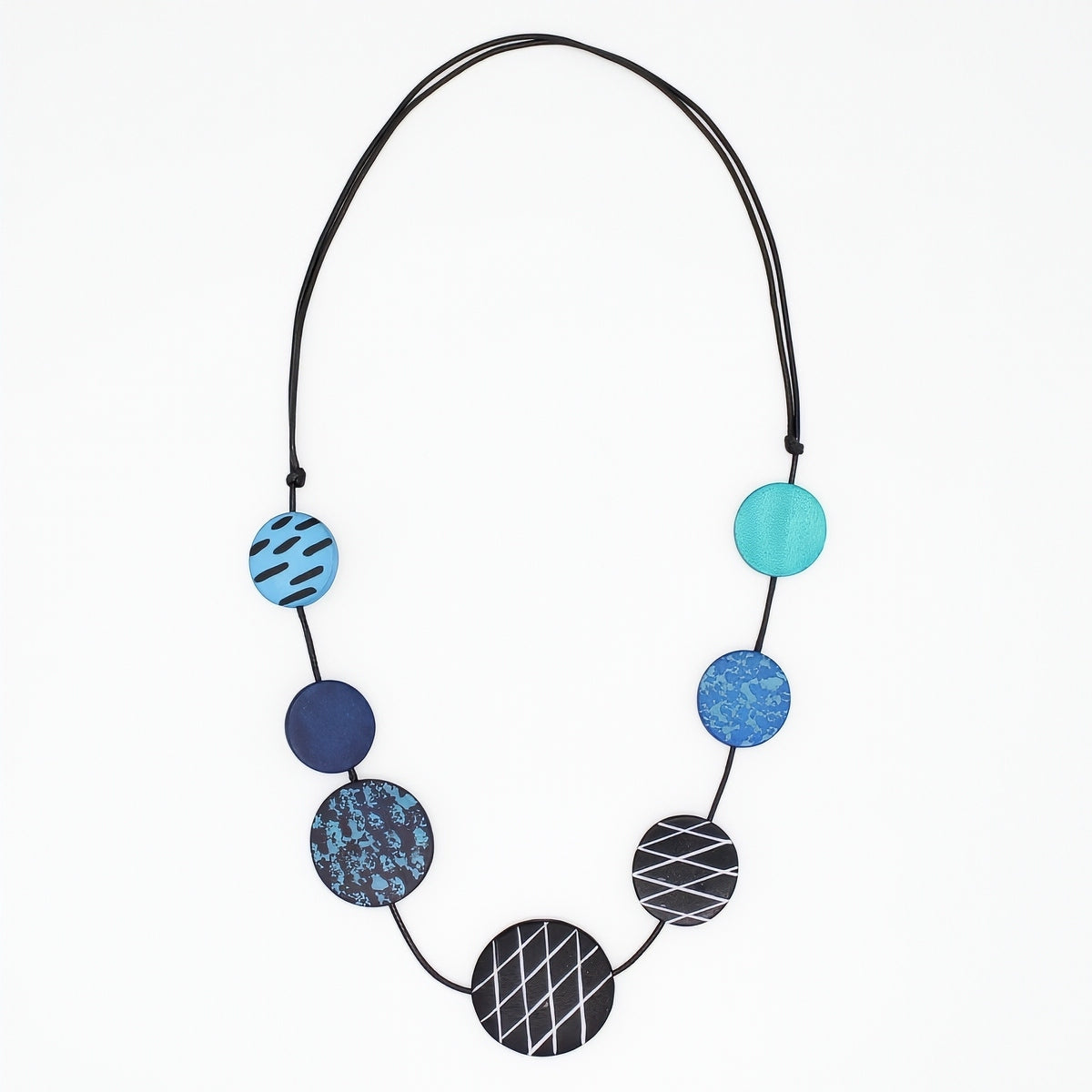 A modern beaded necklace with seven circular discs in shades of blue, teal, and black, featuring various geometric patterns like stripes, grids, and marbled textures, suspended on a black cord against a plain white background.