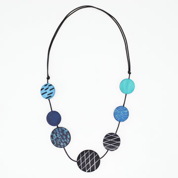 A modern beaded necklace with seven circular discs in shades of blue, teal, and black, featuring various geometric patterns like stripes, grids, and marbled textures, suspended on a black cord against a plain white background.