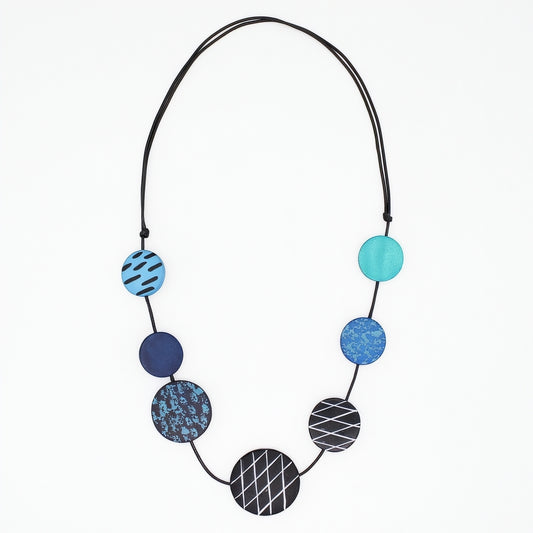 A modern beaded necklace with seven circular discs in shades of blue, teal, and black, featuring various geometric patterns like stripes, grids, and marbled textures, suspended on a black cord against a plain white background.
