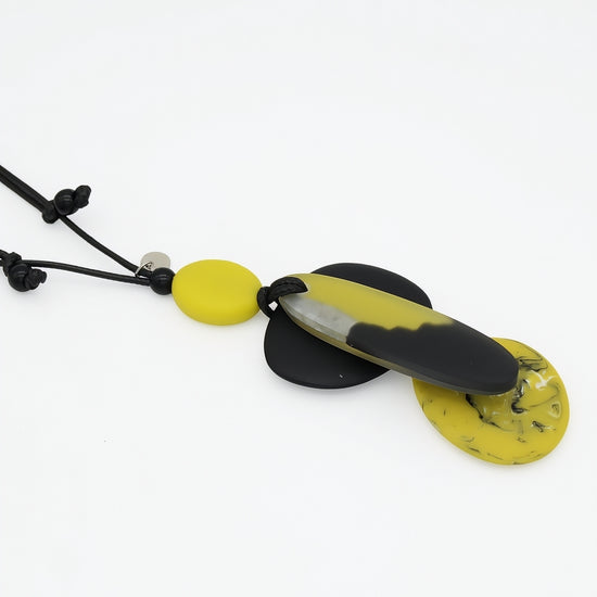 A modern pendant necklace featuring a black cord, a small yellow bead, and a large central pendant with a black and yellow marbled design, flanked by two flat yellow disc pendants on a white background.