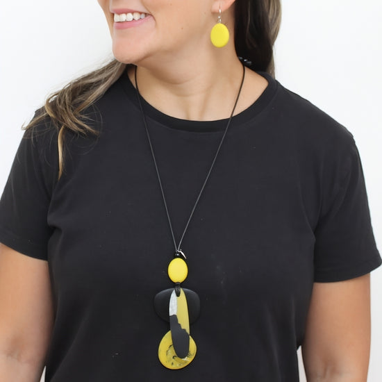 A woman wearing a black t-shirt and a black cord necklace with a yellow and black abstract pendant, matching yellow oval drop earrings, against a plain white background.