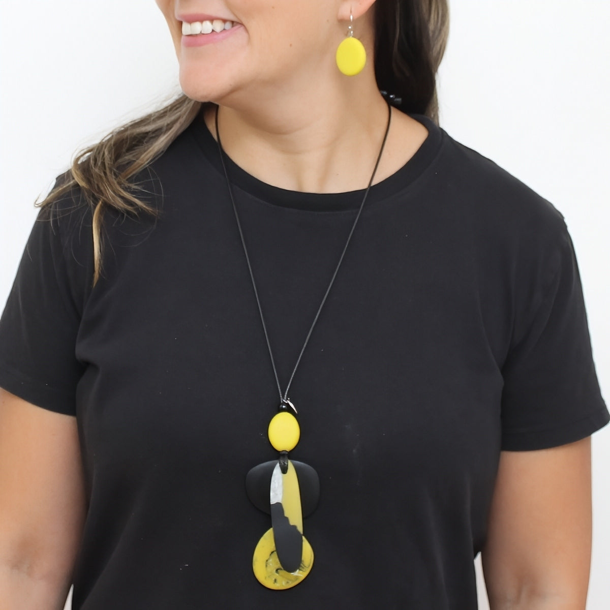 A woman wearing a black t-shirt and a black cord necklace with a yellow and black abstract pendant, matching yellow oval drop earrings, against a plain white background.