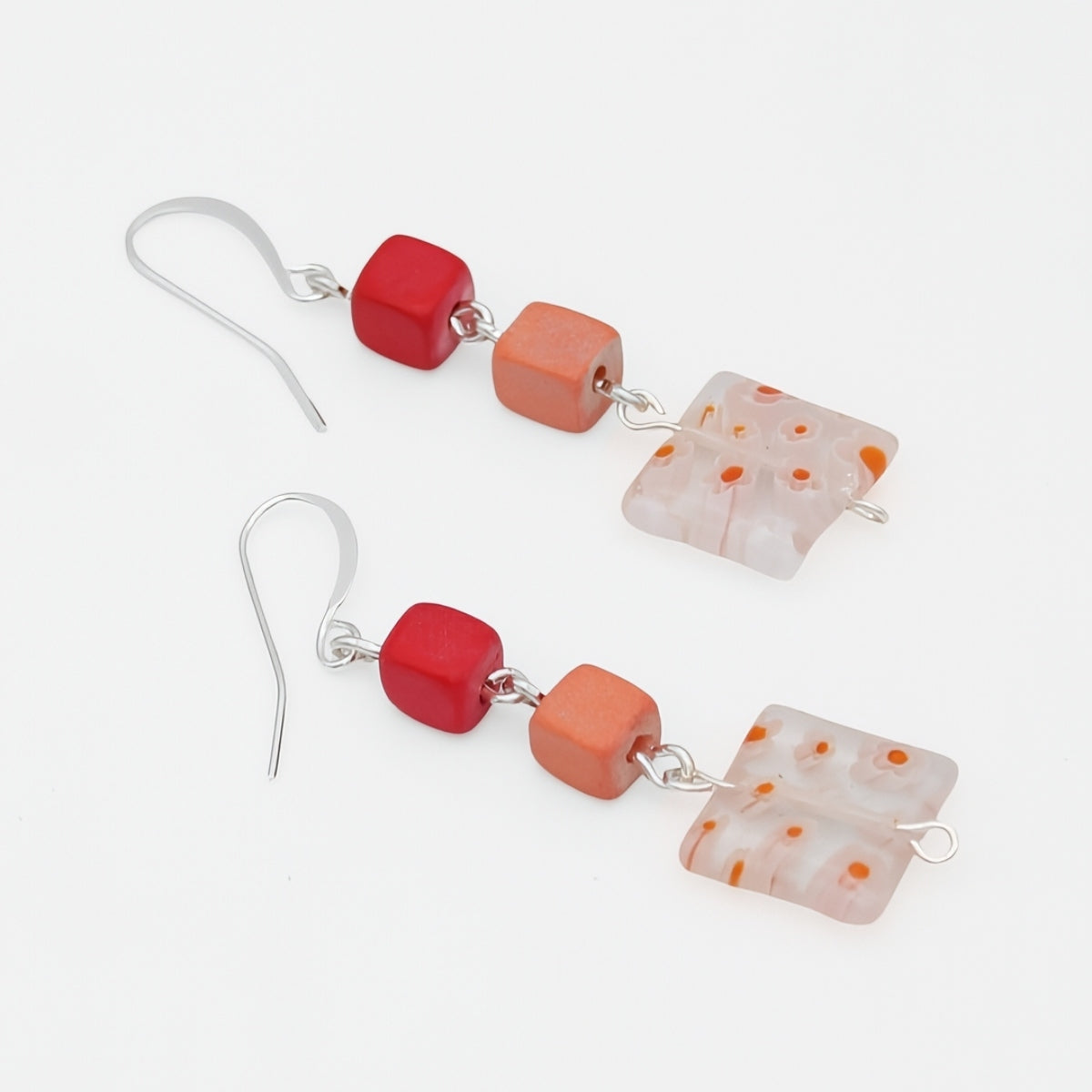 A pair of handmade dangle earrings featuring silver fishhook wires, a glossy red cube bead, a matte orange cube bead, and a translucent white square glass bead with orange speckles, displayed on a clean white background.