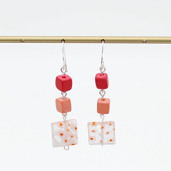 A pair of dangling earrings hanging from a gold rod against a white background. Each earring has a silver fishhook wire, a small red cube bead, a peach-orange cube bead, and a square white glass tile with orange speckles.