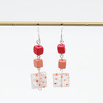 A pair of dangling earrings hanging from a gold rod against a white background. Each earring has a silver fishhook wire, a small red cube bead, a peach-orange cube bead, and a square white glass tile with orange speckles.