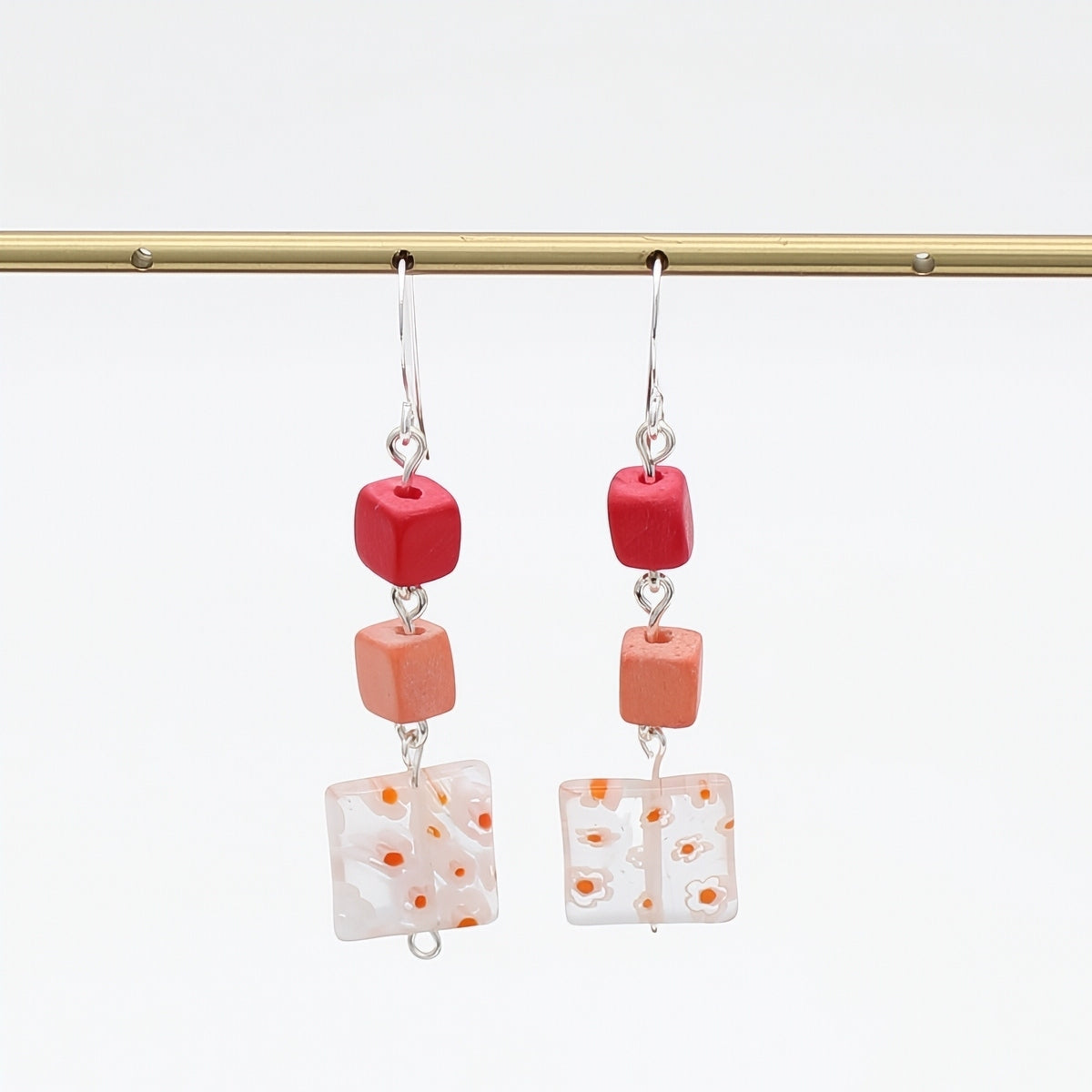 A pair of dangling earrings hanging from a gold rod against a white background. Each earring has a silver fishhook wire, a small red cube bead, a peach-orange cube bead, and a square white glass tile with orange speckles.