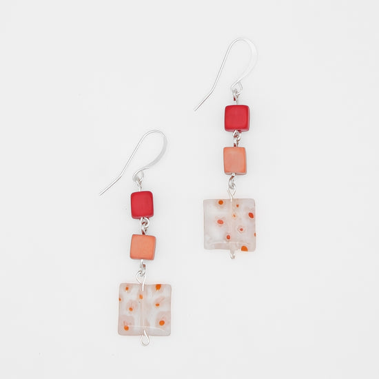A pair of handmade dangle earrings featuring square glass beads in vibrant red, soft orange, and a milky white with orange speckles, suspended from silver fishhook wires on a clean white background.