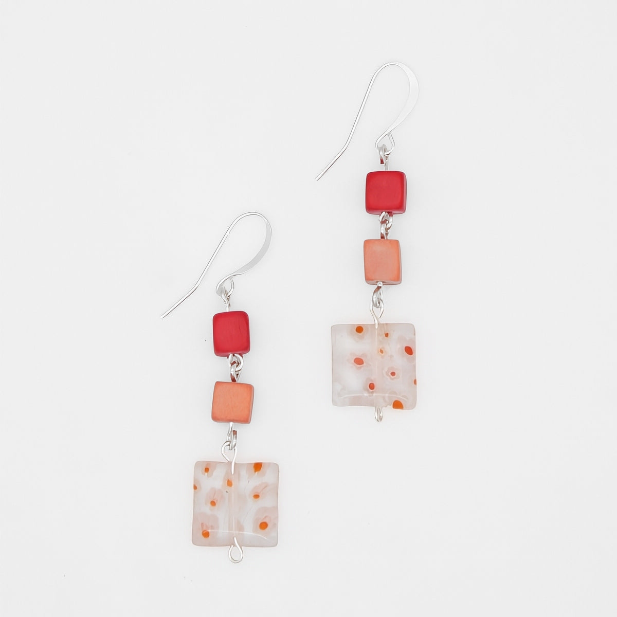 A pair of handmade dangle earrings featuring square glass beads in vibrant red, soft orange, and a milky white with orange speckles, suspended from silver fishhook wires on a clean white background.