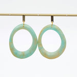 A pair of large, irregularly shaped hoop earrings with a marbled light blue and beige pattern, hanging from gold-toned hooks on a gold display bar against a white background.