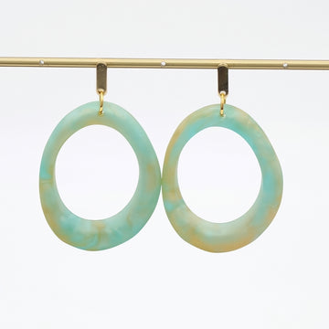 A pair of large, irregularly shaped hoop earrings with a marbled light blue and beige pattern, hanging from gold-toned hooks on a gold display bar against a white background.