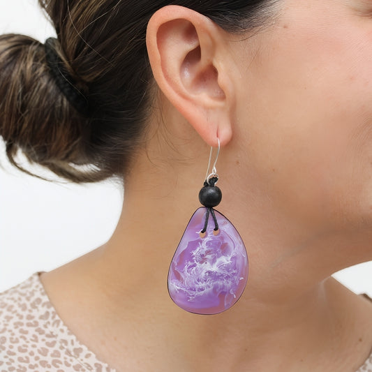 A close-up of a person wearing a teardrop-shaped purple dangle earring with a black bead and silver hook, against a light neutral background.
