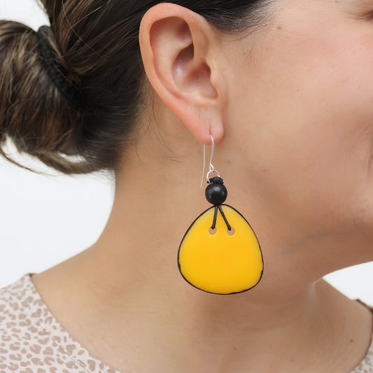 A close-up of a woman's ear wearing a dangling earring. The earring has a bright yellow teardrop-shaped drop with black hand-painted outlines and a small black bead above it on a silver hook.