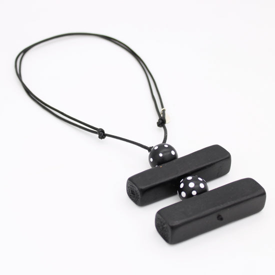 Black wooden pendant necklace with two rectangular bars connected by a black cord and accented with two black beads featuring white polka dots, shown on a plain white background.