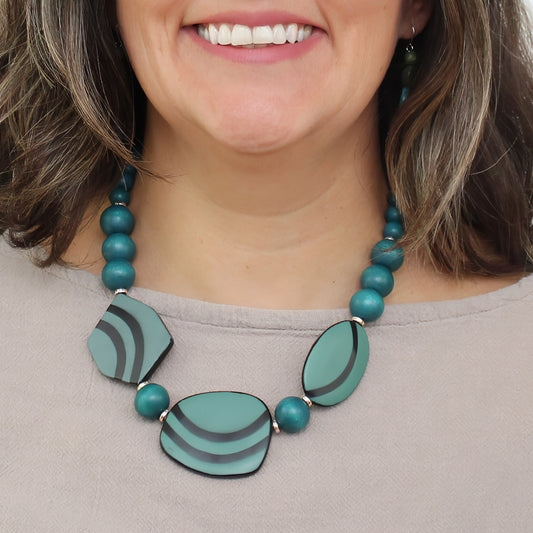 A close-up of a woman wearing a statement necklace with teal and mint green marbled stone-like beads and matching teal beads, paired with a taupe top.